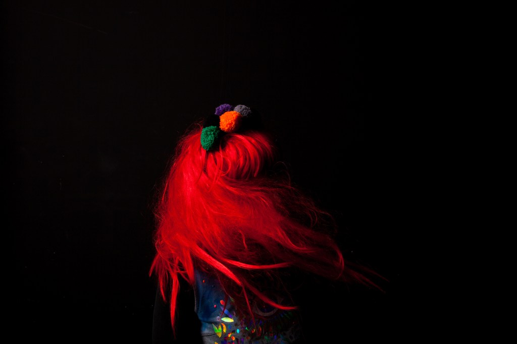 A black background, a body with bright red hair and a costume whirls. We can't see the person's face, just the vibrant red of a wig and a pom pom headband.