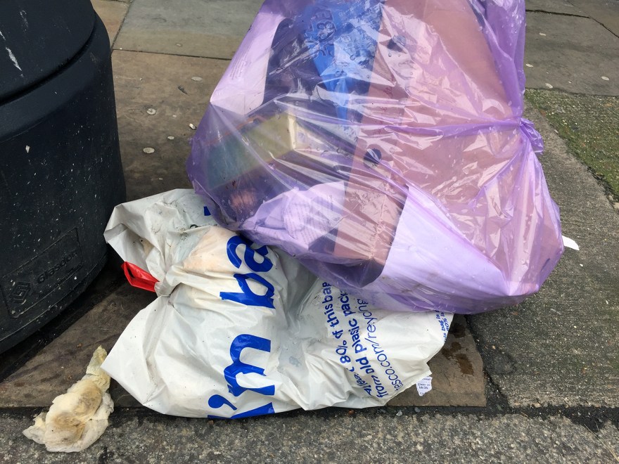 A tesco bag, tied at the top with the words 'I'm back' partially visible, it is squashed by a larger lavender trashbag.